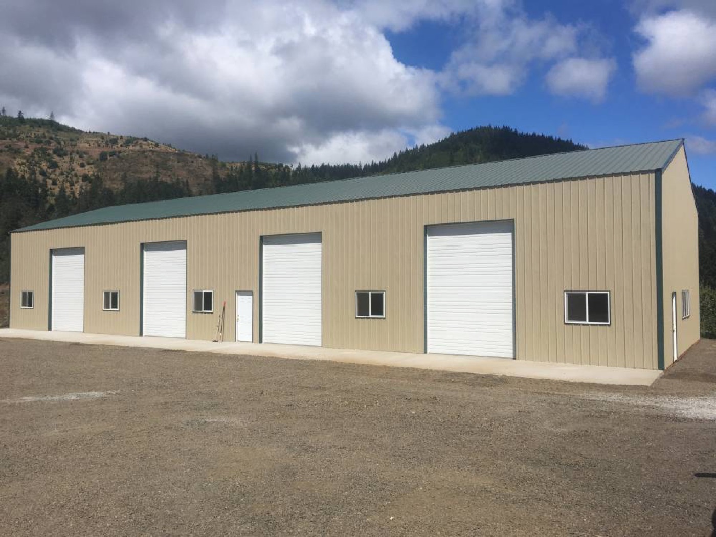 Tan shop with a green roof and four roll-up doors against a mountain backdrop, built by Miner Pole Buildings