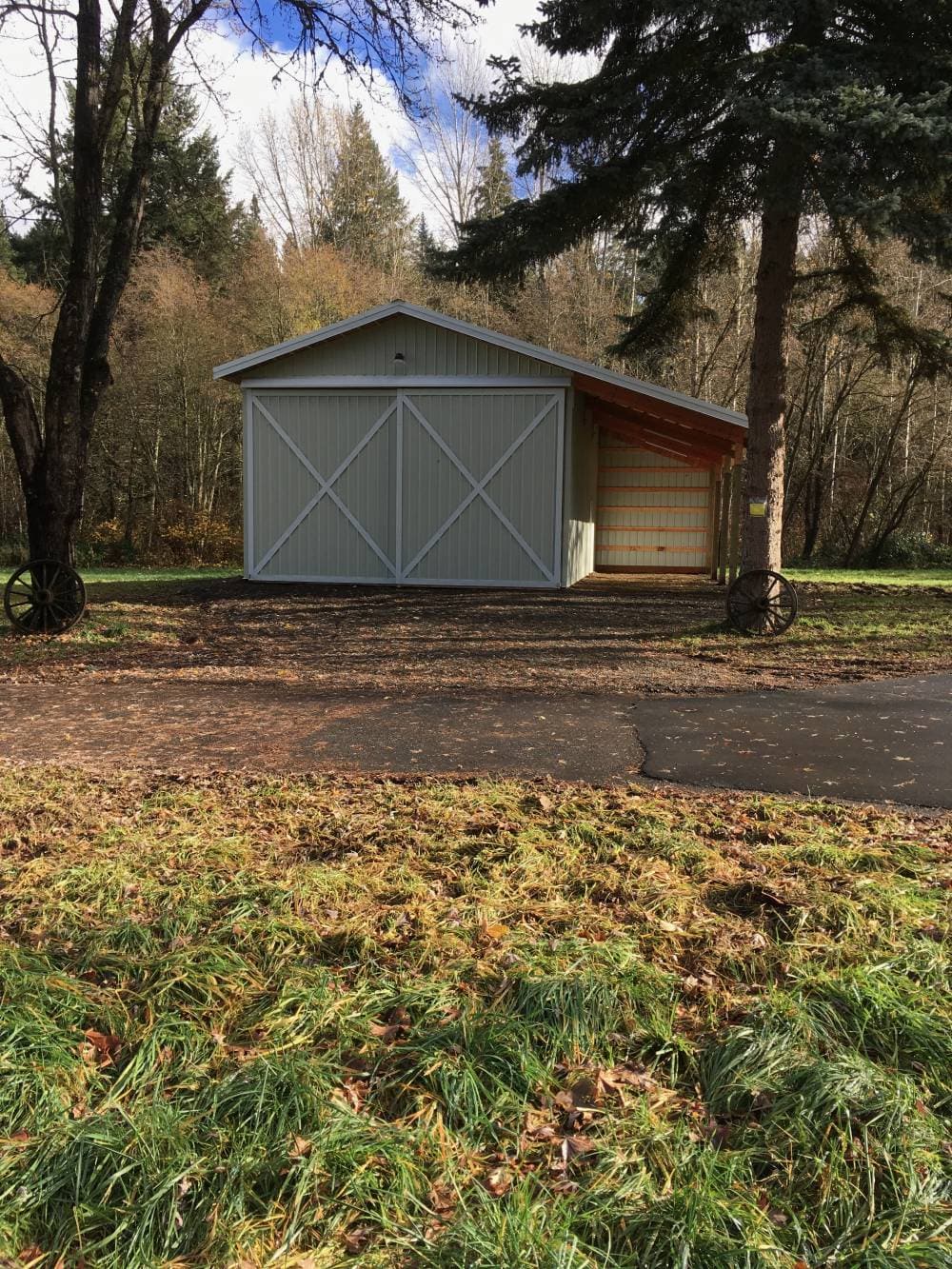 Sage-green gable post-frame barn with white X-doors and a lean-to on a wooded property, built by Miner Pole Buildings